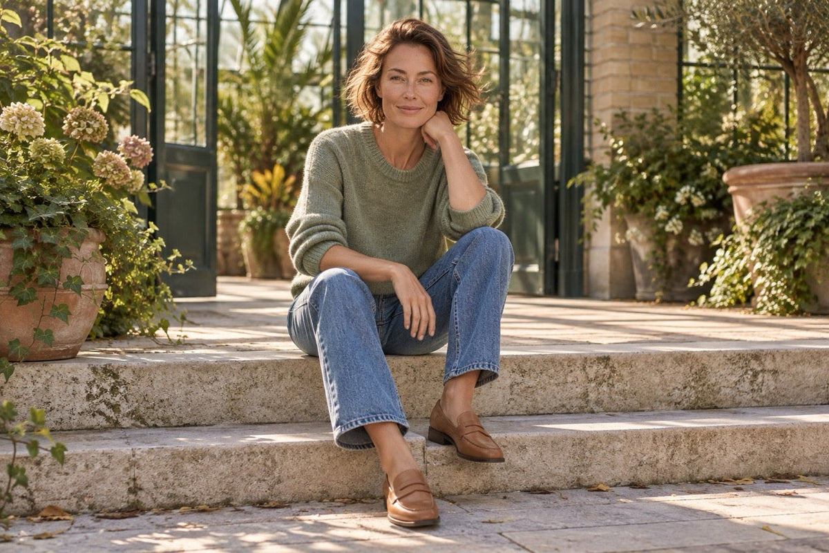 Woman wearing a sage-green jumper, blue jeans and tan loafers, sitting on stone steps outside a sunlit conservatory.